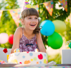 Little girl sitting in front of a birthday cake and smiling, wearing a birthday hat with balloons in background